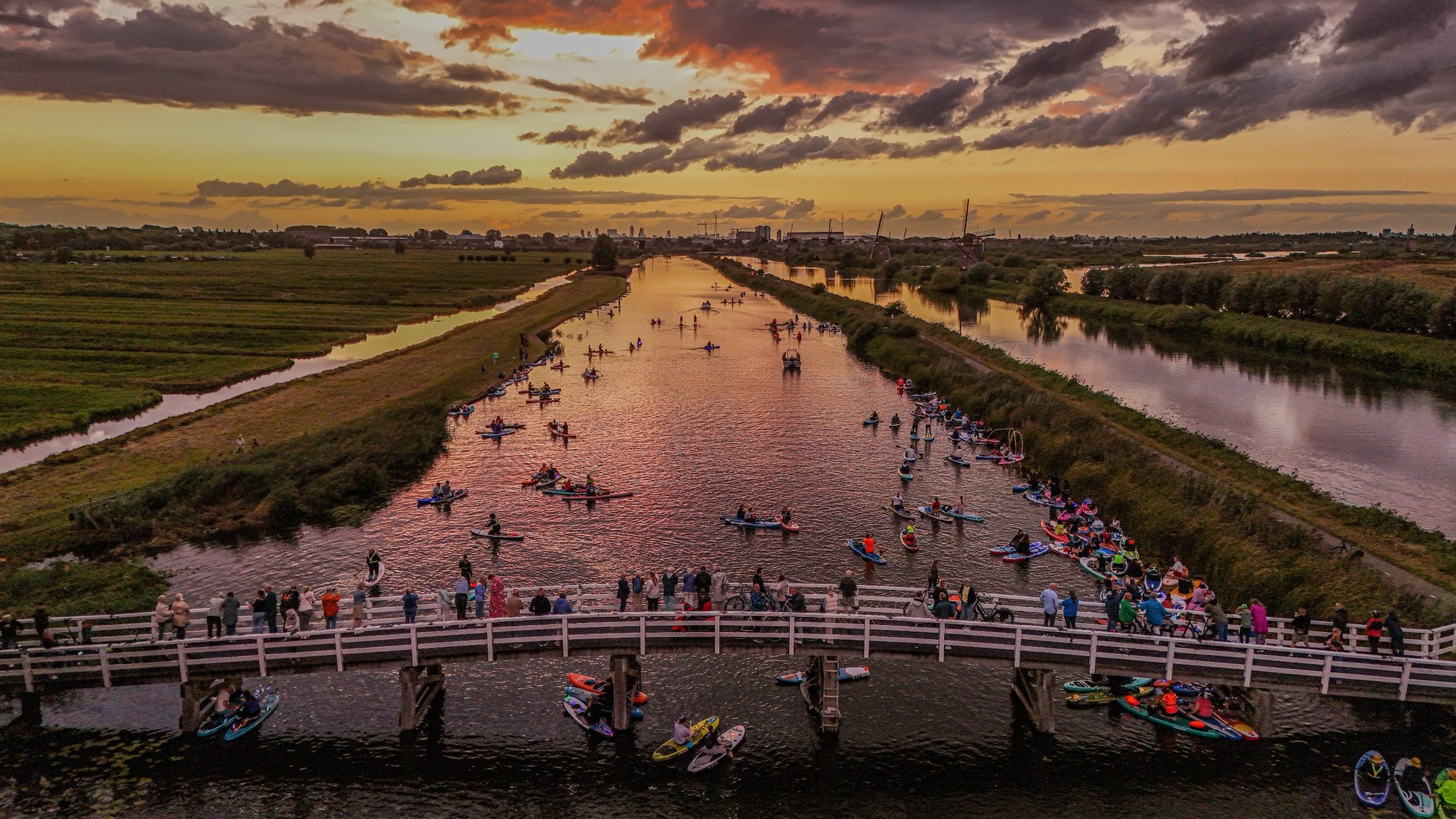 Mensen op kajaks en paddleboards op een kanaal bij zonsondergang met toeschouwers op een houten brug.