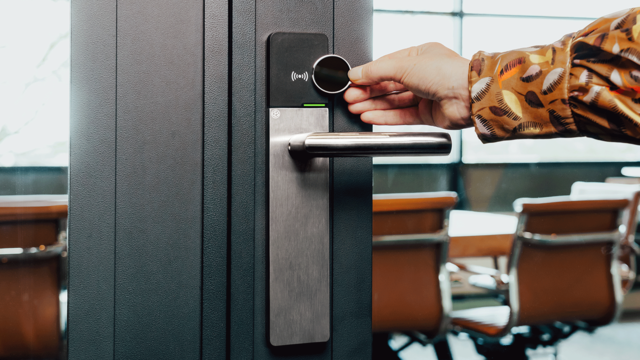 Hand houdt een ronde transponder tegen een elektronisch deurslot op een grijze deur, met kantoorstoelen zichtbaar door het glas.