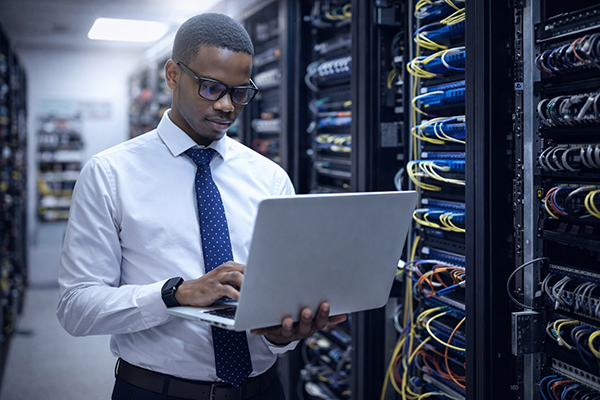 Man in een witte overhemd en stropdas die met een laptop werkt in een serverruimte met kabels en servers.