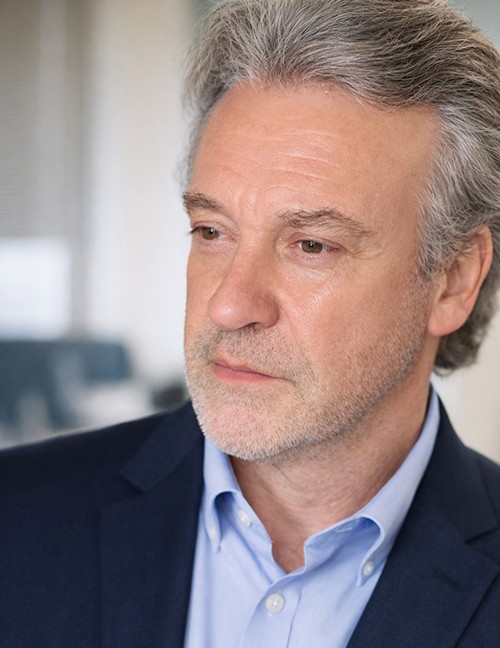 Portrait van een oudere man met grijs haar en stoppelbaard, gekleed in een blauw overhemd en donkerblauw colbert.