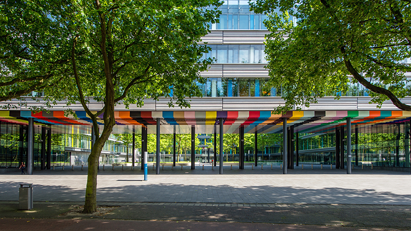 Moderne glazen kantoorgebouw met een kleurrijke overkapping en bomen op de voorgrond.