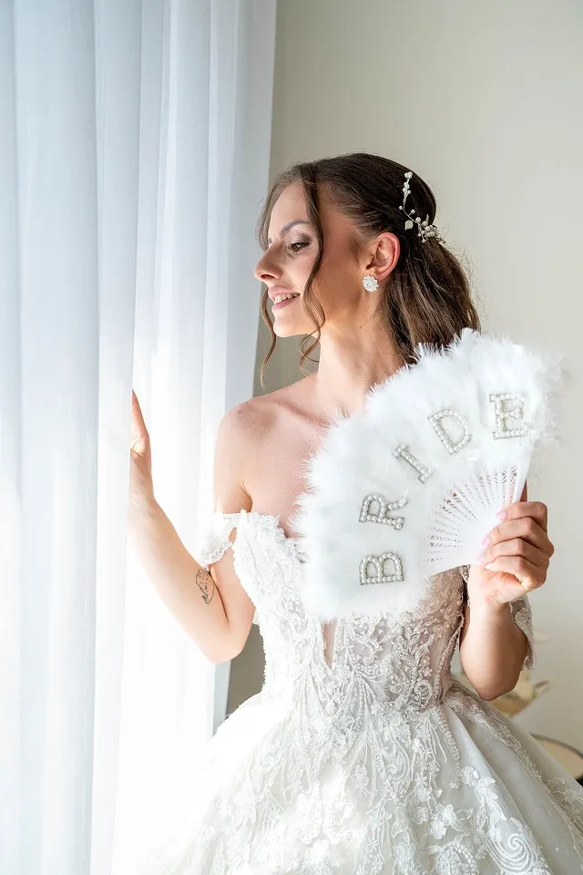 Mlada u čipkanoj venčanici pozira kraj prozora sa belom lepezom.