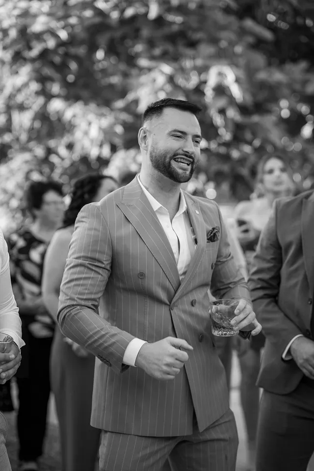 Nasmejan muškarac u elegantnom odelu tokom venčanja u Leskovcu – spontani crno-beli dokumentarni trenutak.