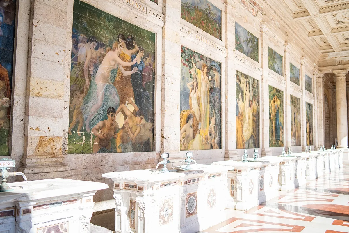 Prostrana galerija sa zidnim mozaicima i mermernim fontanama u istorijskom objektu u Italiji.