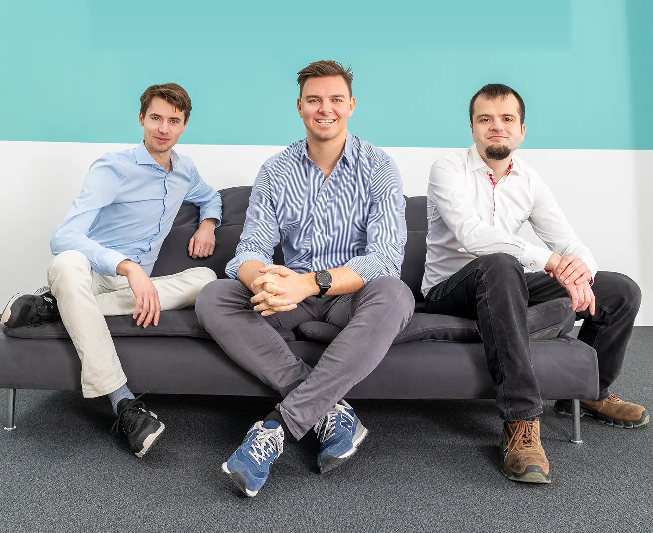 Three men sitting on a dark gray sofa against a blue and white wall, smiling and facing the camera.