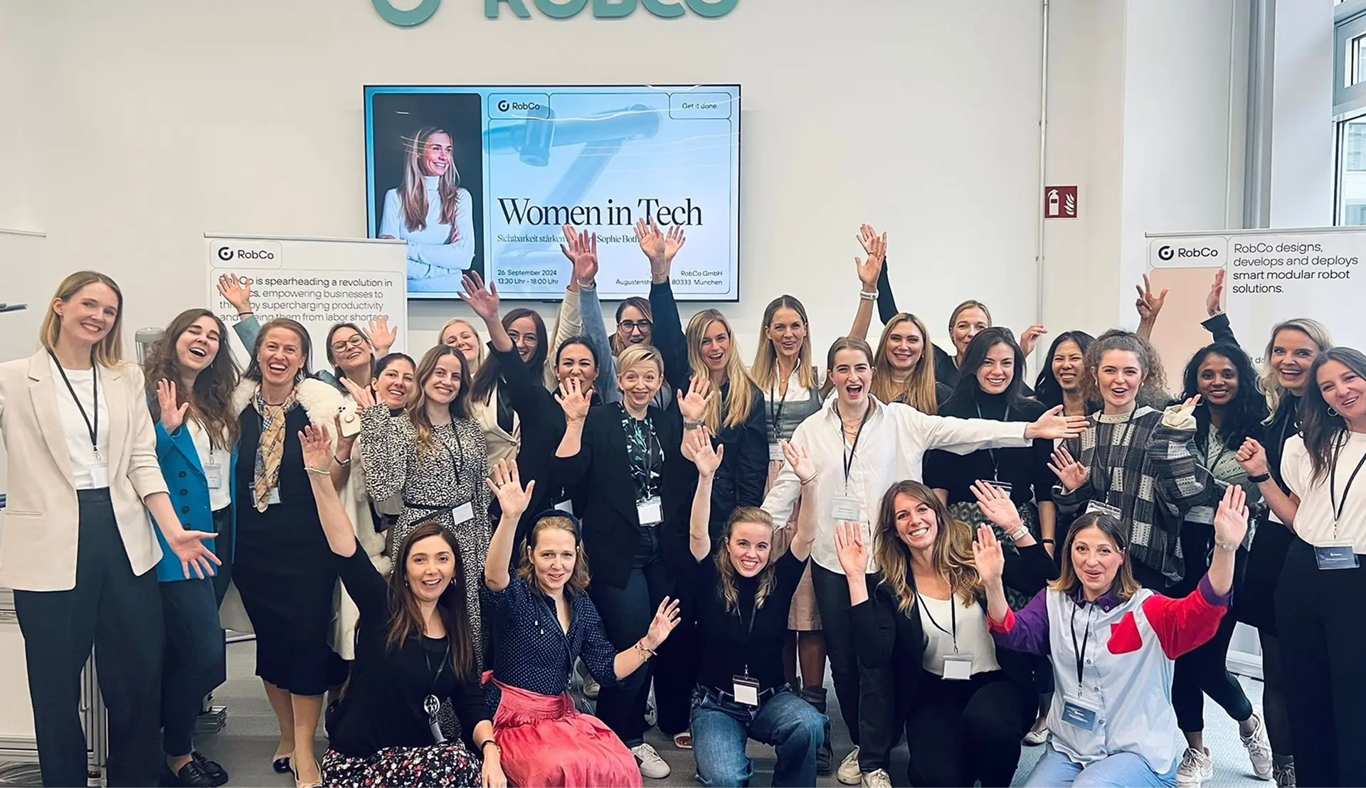Group of smiling women posing together indoors with raised hands in front of a screen displaying 'Women in Tech' event information.