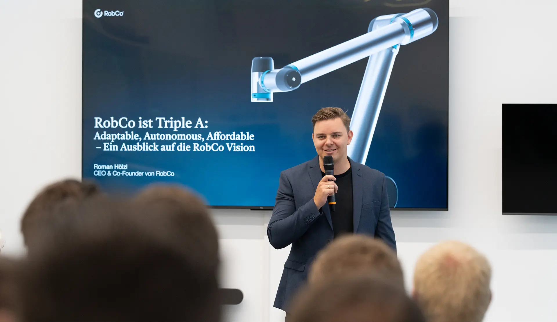 Man in a dark suit speaking into a microphone in front of a screen displaying a RobCo robotic arm and presentation text.