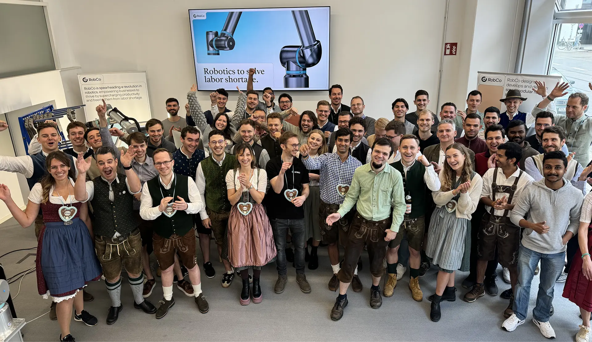 Large group of people in traditional Bavarian clothing and casual attire posing indoors in front of screens displaying robotics messages.