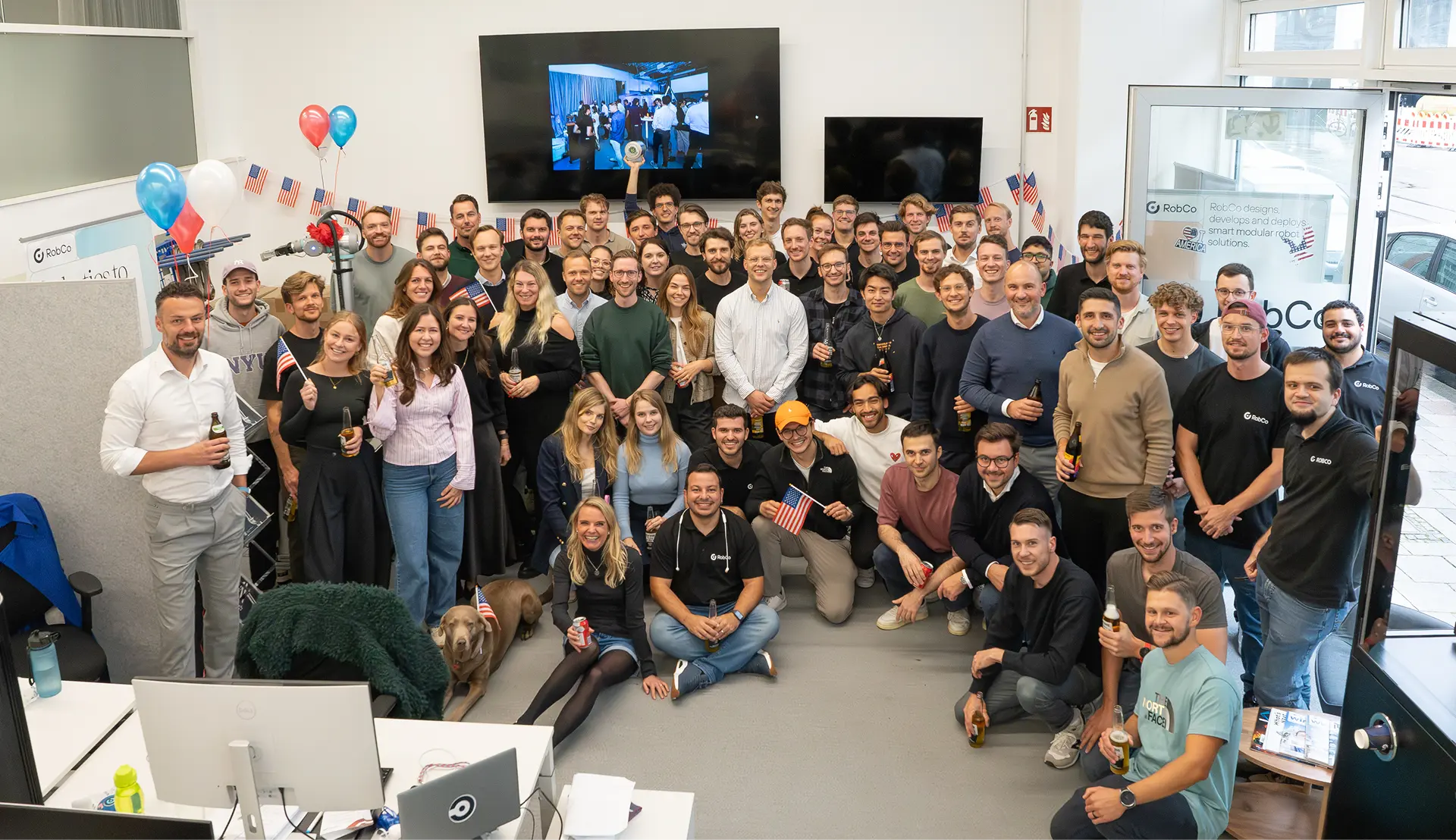 Large group of diverse people posing and smiling in a decorated office with American flags and balloons.