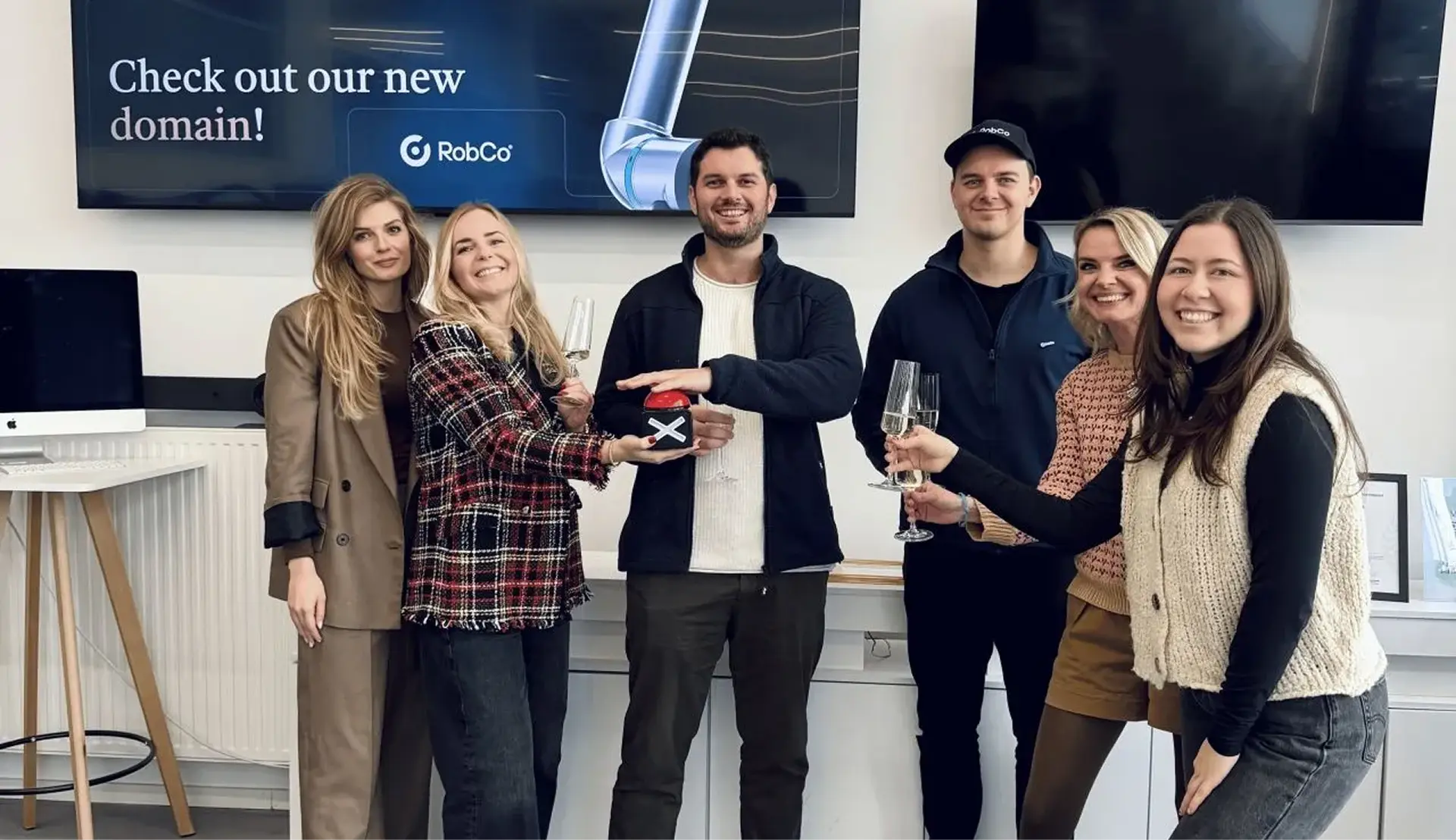 Six smiling people holding champagne glasses and a red button, standing in front of monitors with text promoting a new domain.