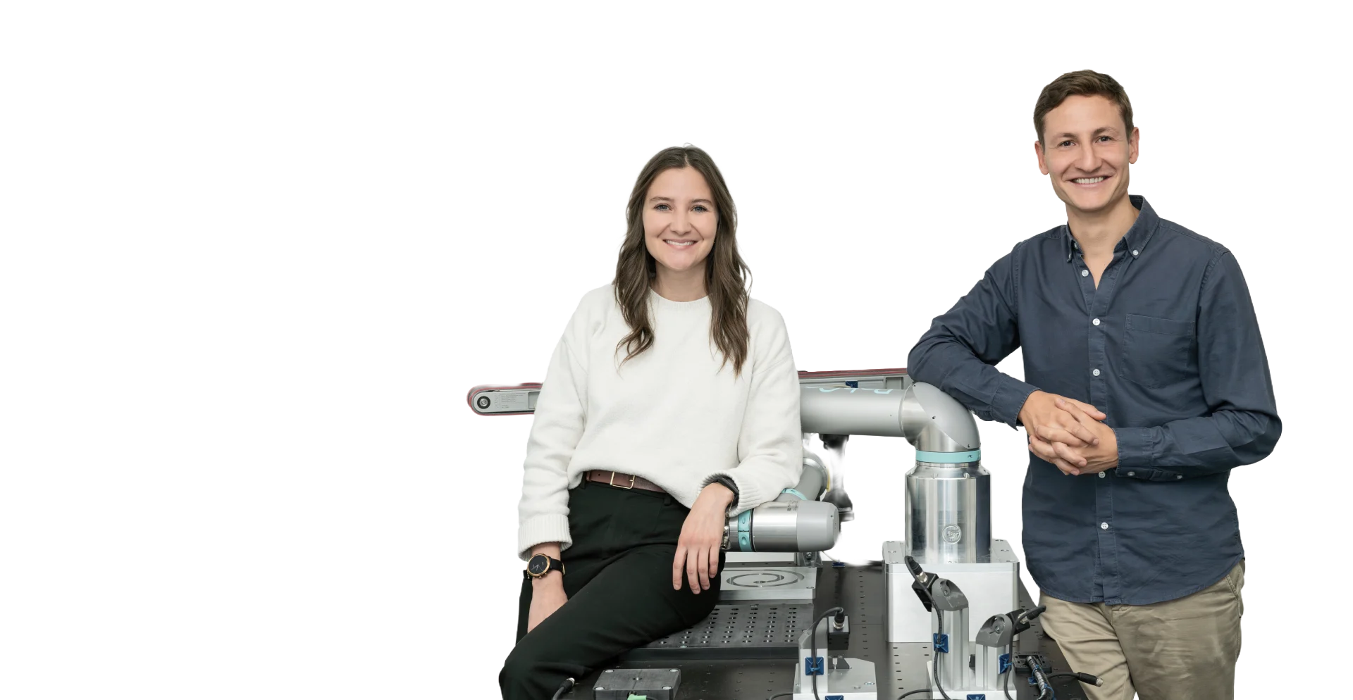 Smiling woman in white sweater and man in blue shirt standing by a robotic arm on a workbench.