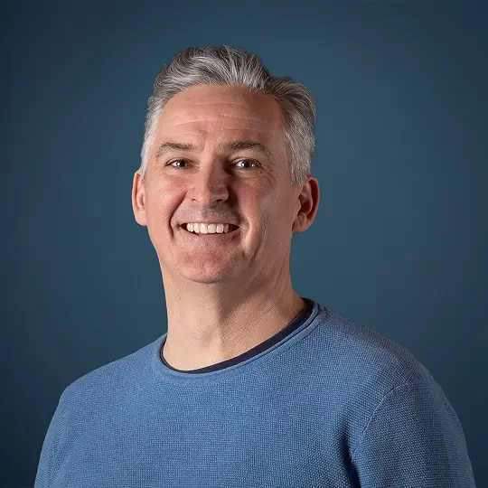 Smiling person with short blond hair and hoop earrings wearing a textured jacket.