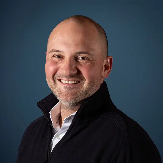 Smiling person with short blond hair and hoop earrings wearing a textured jacket.