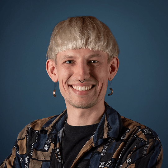 Smiling person with short blond hair and hoop earrings wearing a textured jacket.