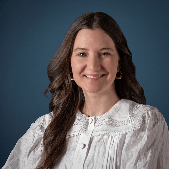 Smiling woman with wavy brown hair wearing a black turtleneck sweater and gold hoop earrings against a plain white background.