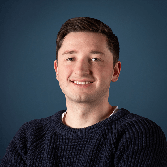 Portrait of a smiling young man with light skin, short brown hair, wearing a dark zip-up sweater over a light shirt.