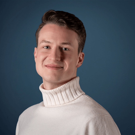 Portrait of a smiling young man with short brown hair wearing a white shirt against a plain light background.