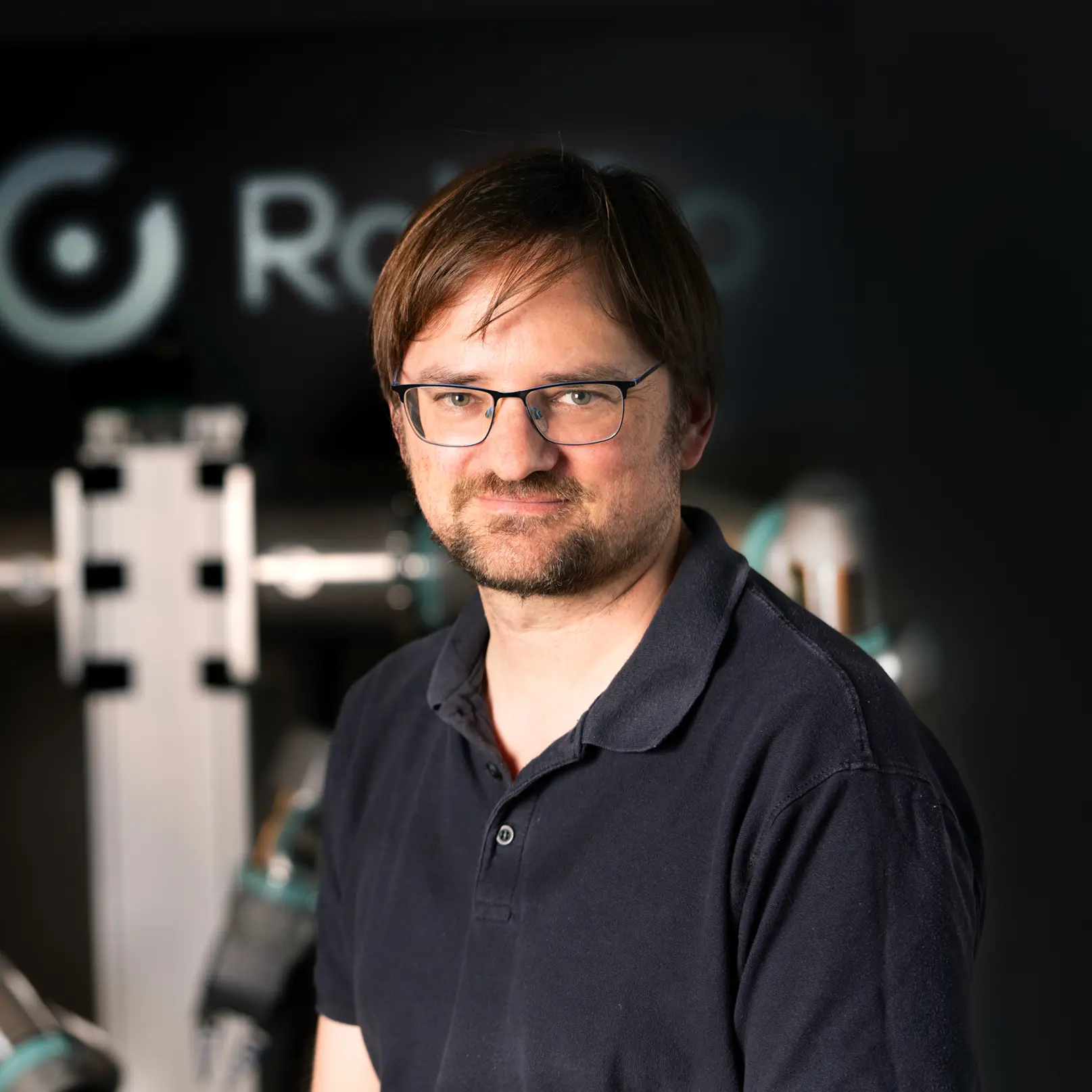 Man with glasses and short brown hair wearing a dark polo shirt, standing in front of robotic equipment with a blurred RobCo logo in the background.