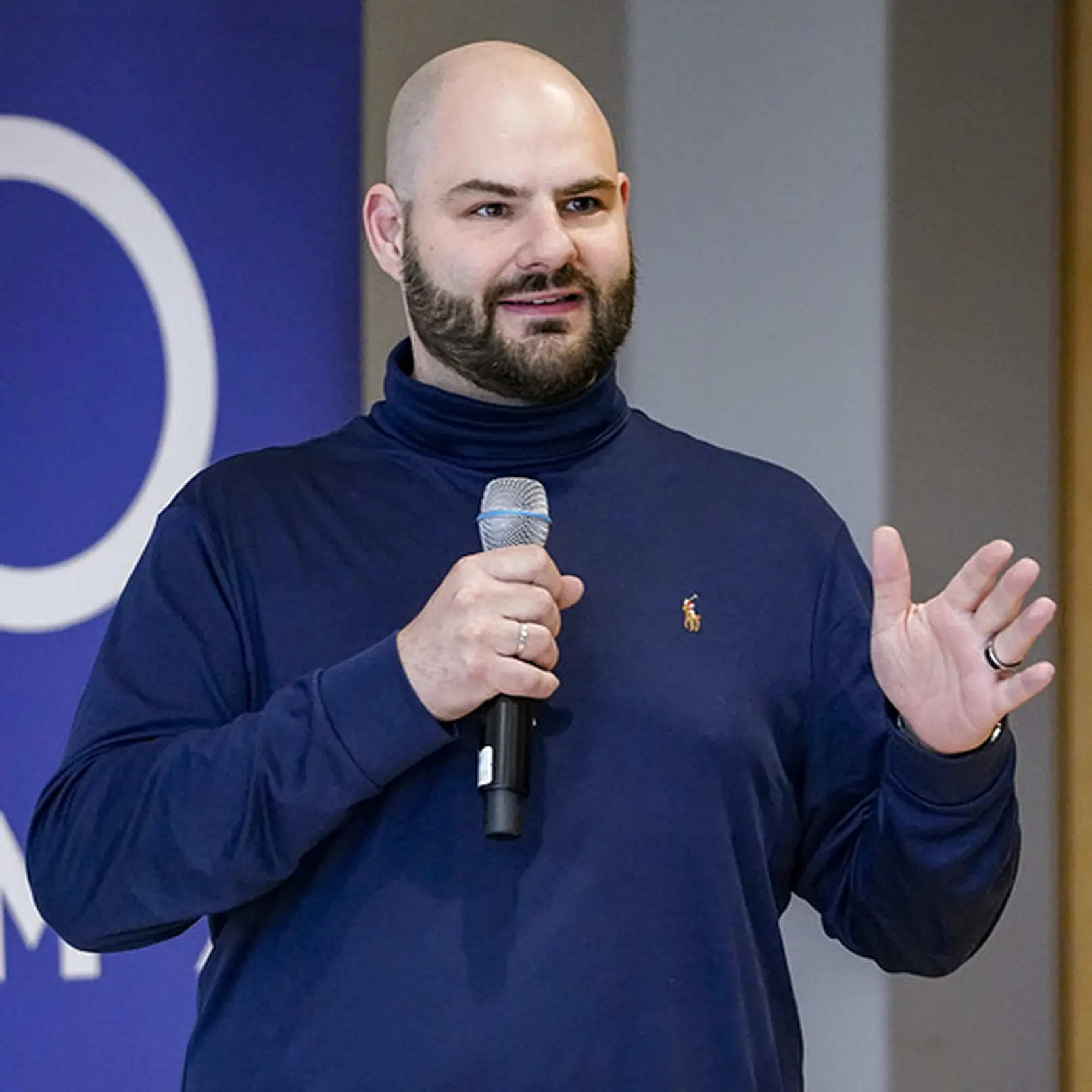 Bearded man in a navy blue turtleneck speaking into a microphone with a blue background.