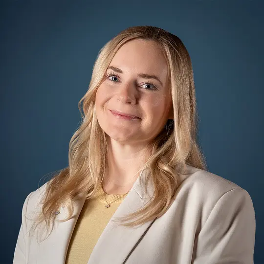 Smiling person with short blond hair and hoop earrings wearing a textured jacket.