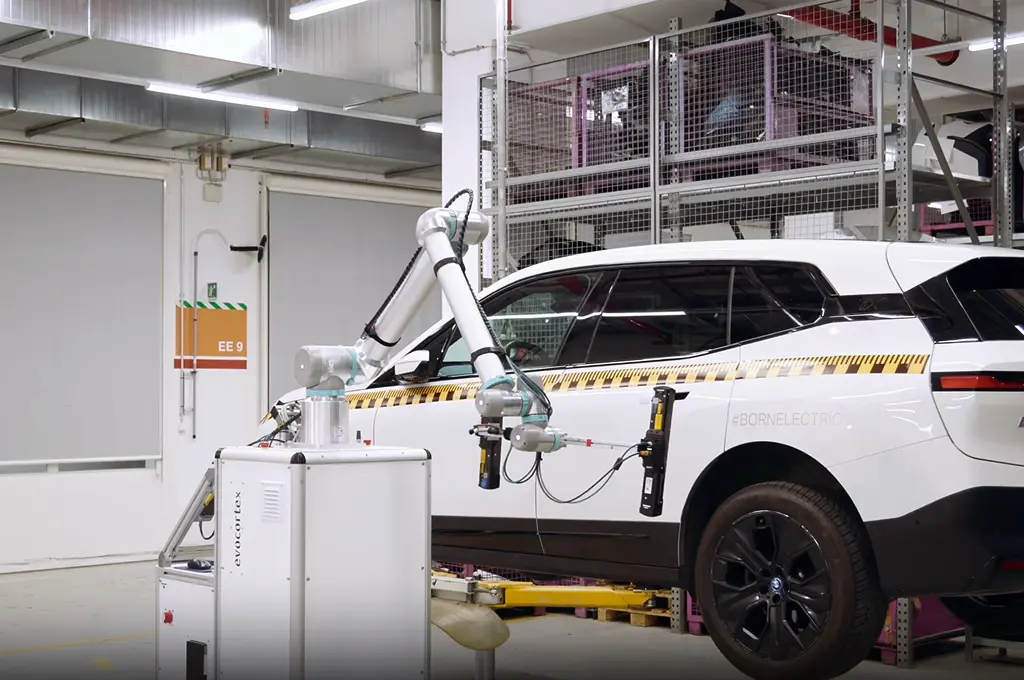 Robotic arm conducting a test on the side of a white electric SUV in an industrial facility.