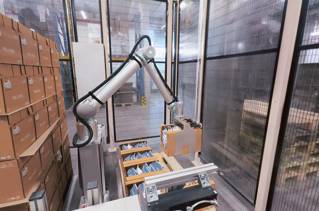 Robotic arm picking a cardboard box in an automated warehouse storage area with stacked boxes.