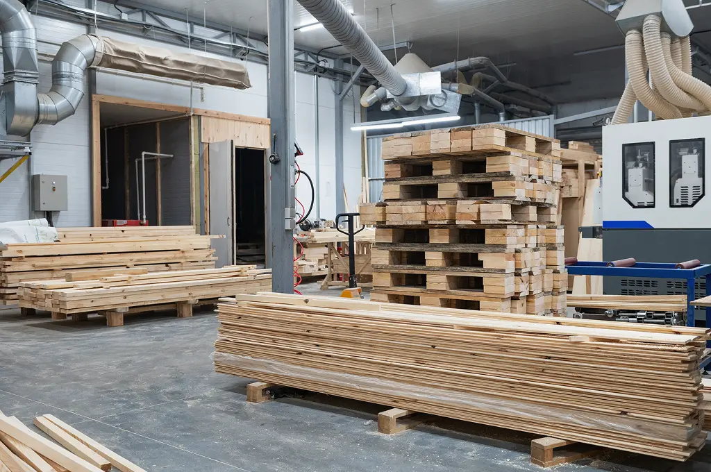 Stacks of cut wooden planks and pallets in a large industrial workshop with machinery and ventilation pipes.