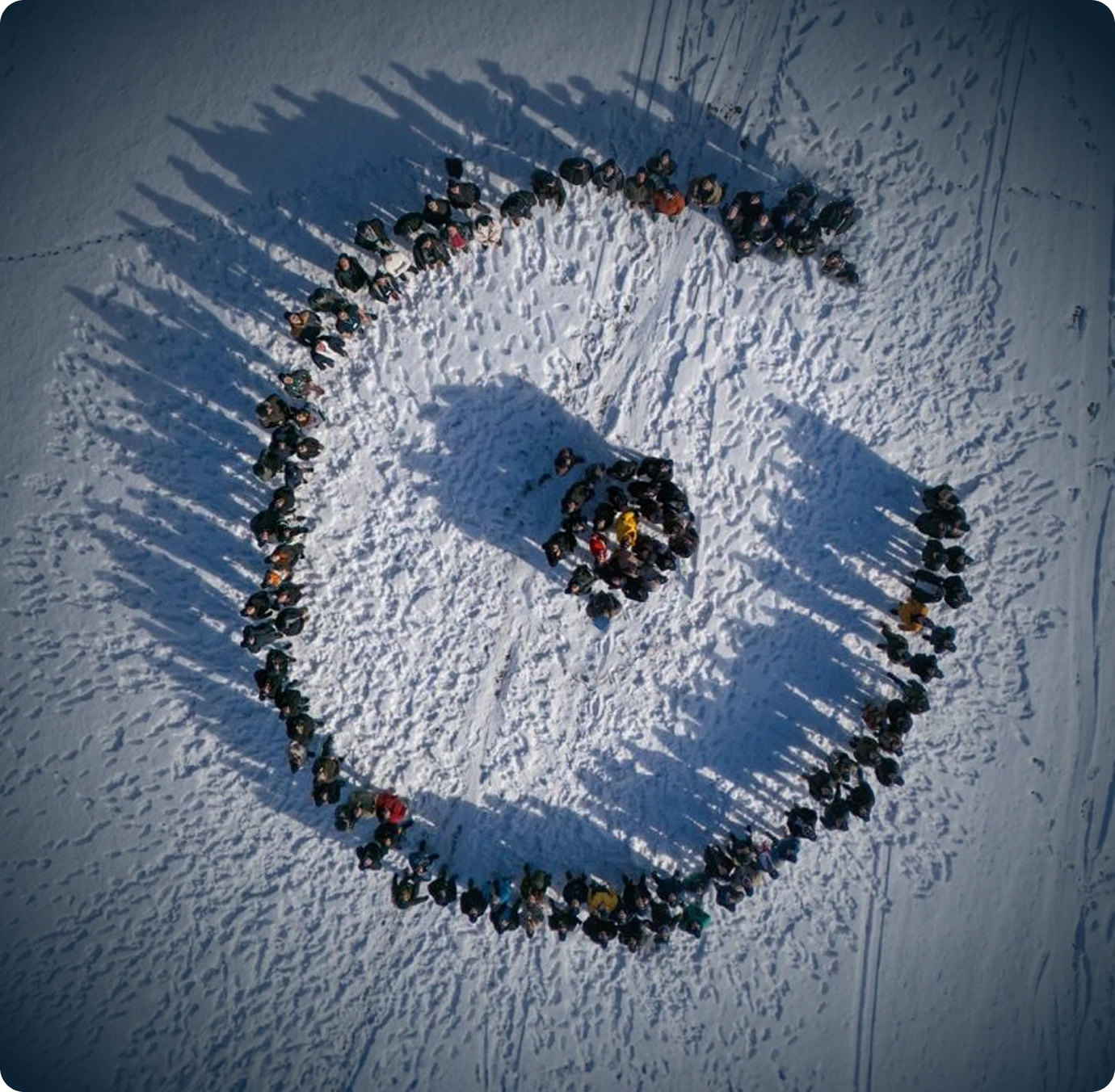 Aerial view of people standing in a large circle on snow, shadows extending outward.