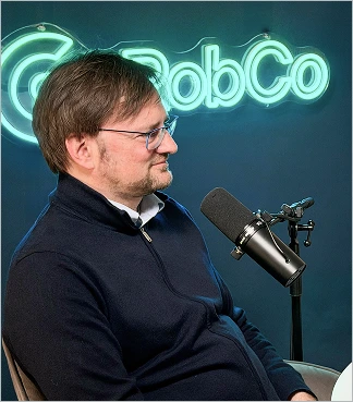 Man wearing glasses speaking into a microphone with illuminated 'RobCo' sign behind him.