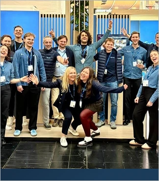 Group of people smiling and posing energetically indoors with blue panels behind them.