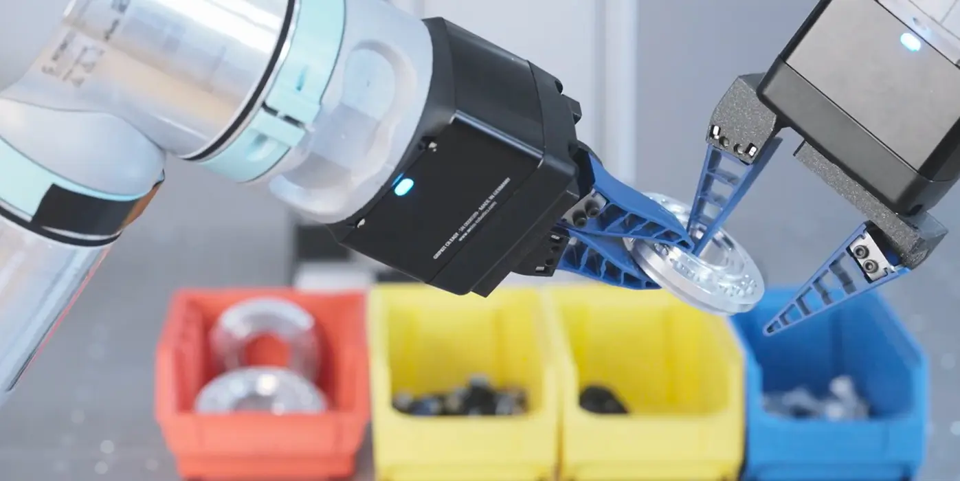 Robotic arm with blue mechanical gripper holding a clear round plastic or glass component above red, yellow, and blue bins containing various small parts.