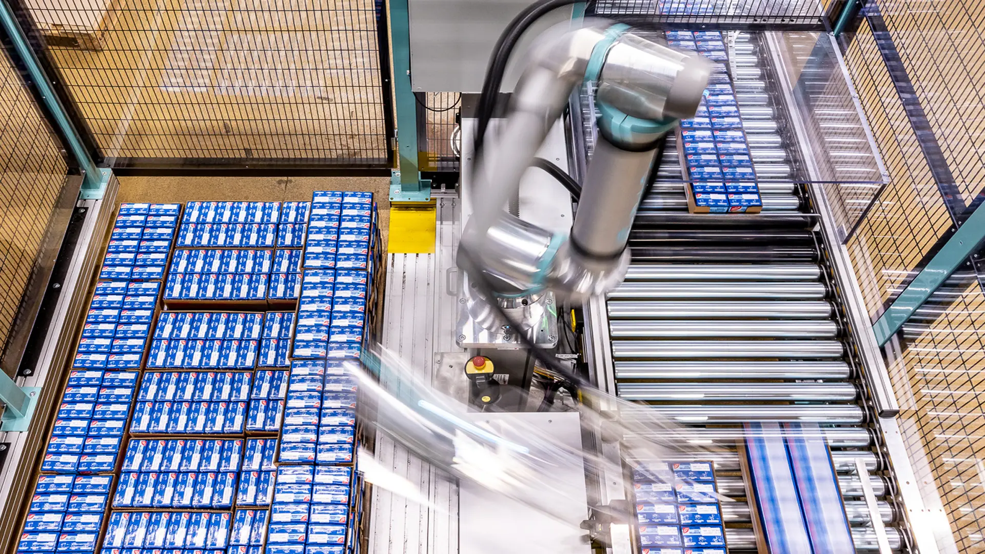 Industrial robot arm rapidly moving on a conveyor belt system sorting or packing numerous blue and white boxed products inside a fenced manufacturing area.