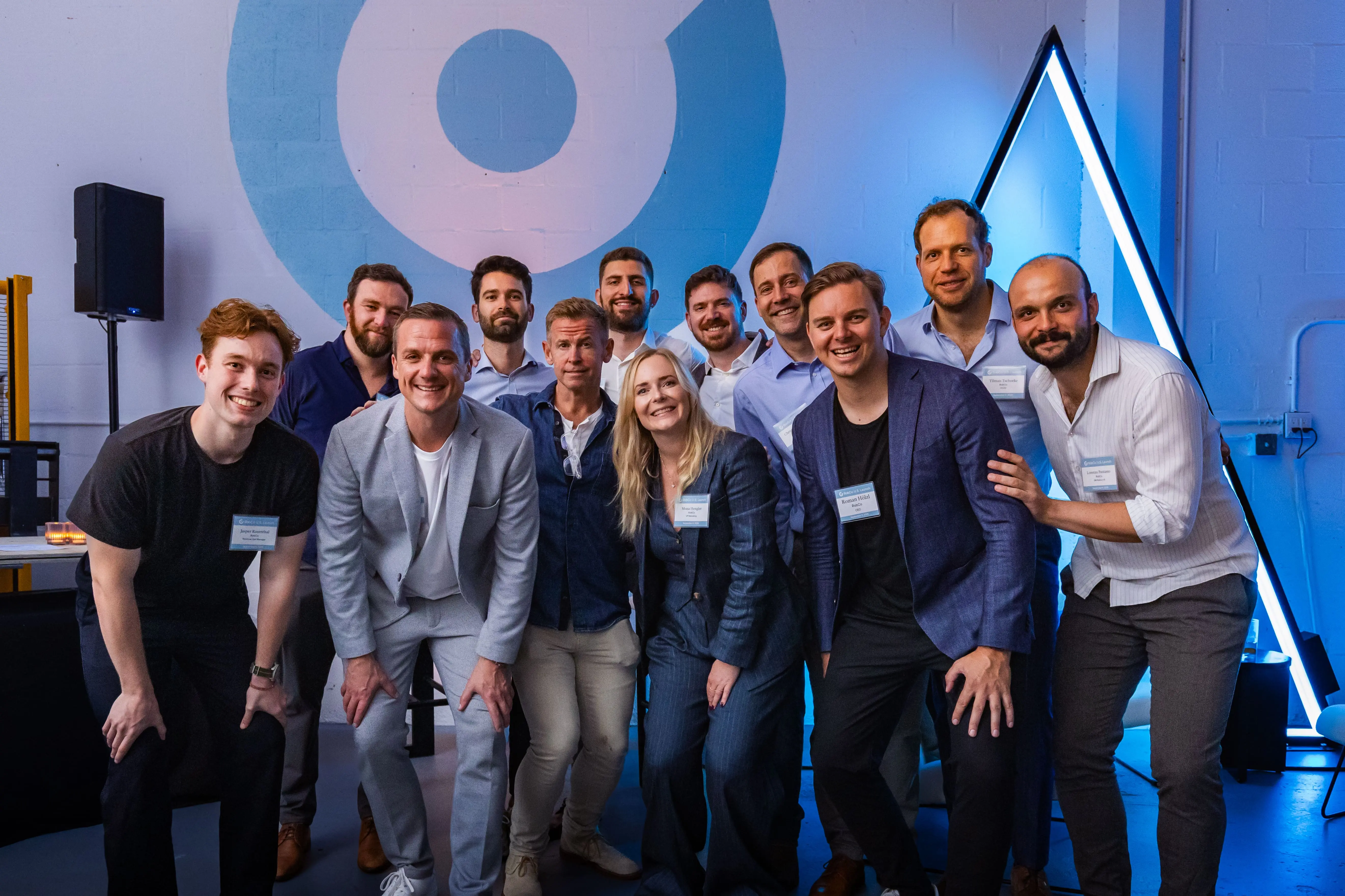 Group of twelve people at RobCo’s US Launch Office Opening. Eleven men and one woman are smiling and posing for a group photo in a modern, brightly lit space with a blue and white wall graphic and a glowing triangle in the background.