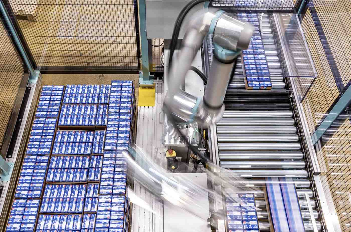 Ein Roboterarm verschiebt mit präziser Kinematik Kartons mit blauen Verpackungen auf einem Förderband in einer Produktionshalle, wobei die fließenden Bewegungen des Arms im Einsatz zu sehen sind.