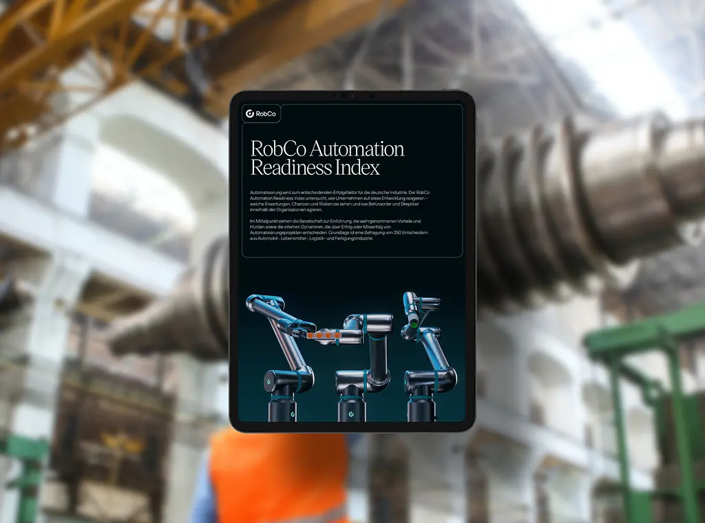 Ein Tablet zeigt den „RobCo Automation Readiness Index“ mit einem Titeltext und drei modularen Roboterarmen auf dunklem Hintergrund. Im unscharfen Hintergrund ist eine industrielle Produktionshalle mit einer Person in Warnweste zu sehen.