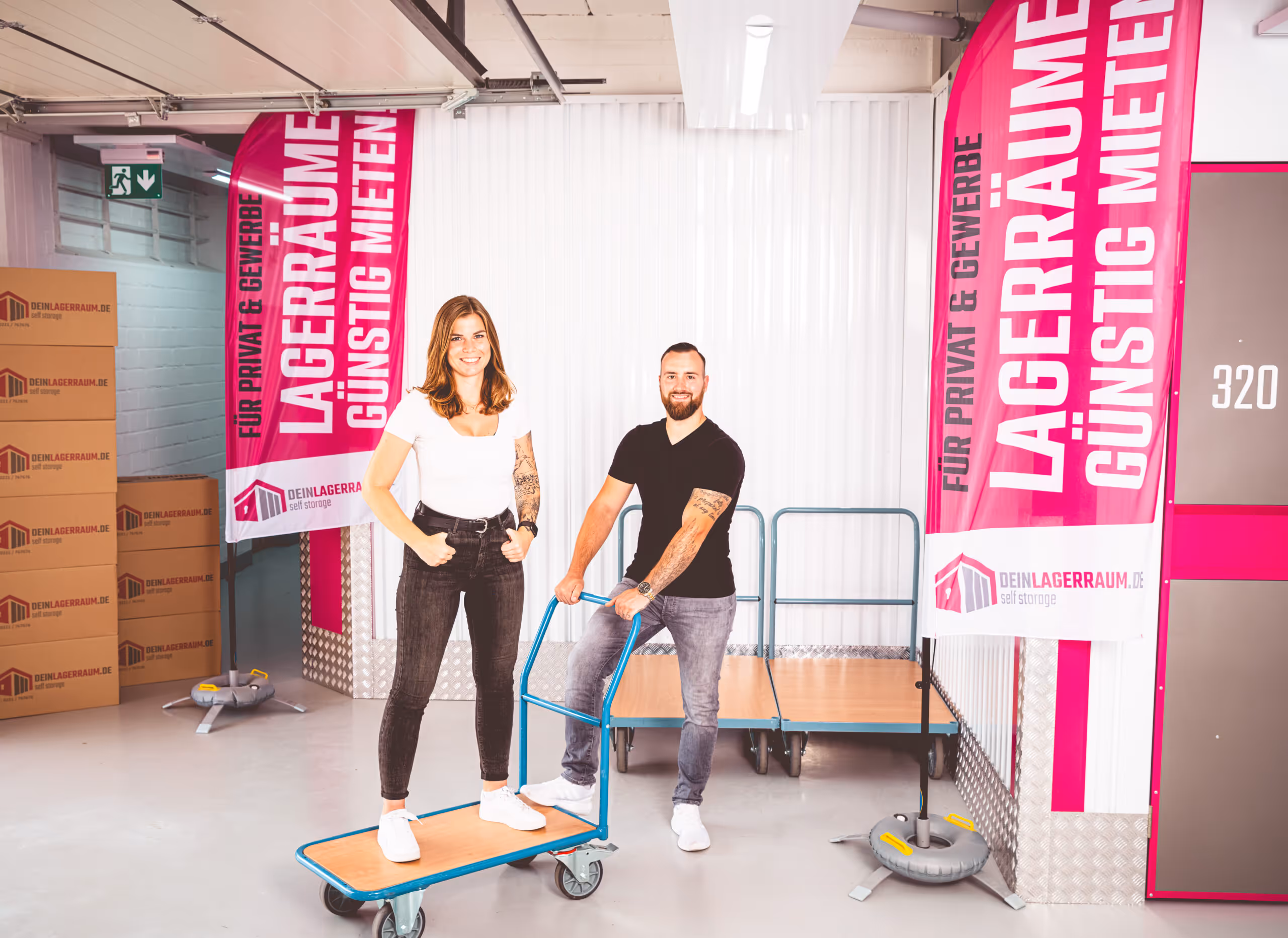 Zwei Personen in einem Lagerraum stehen lachend auf einem Möbelroller, umgeben von pinken Bannern mit Werbung für günstige Lagerräume.