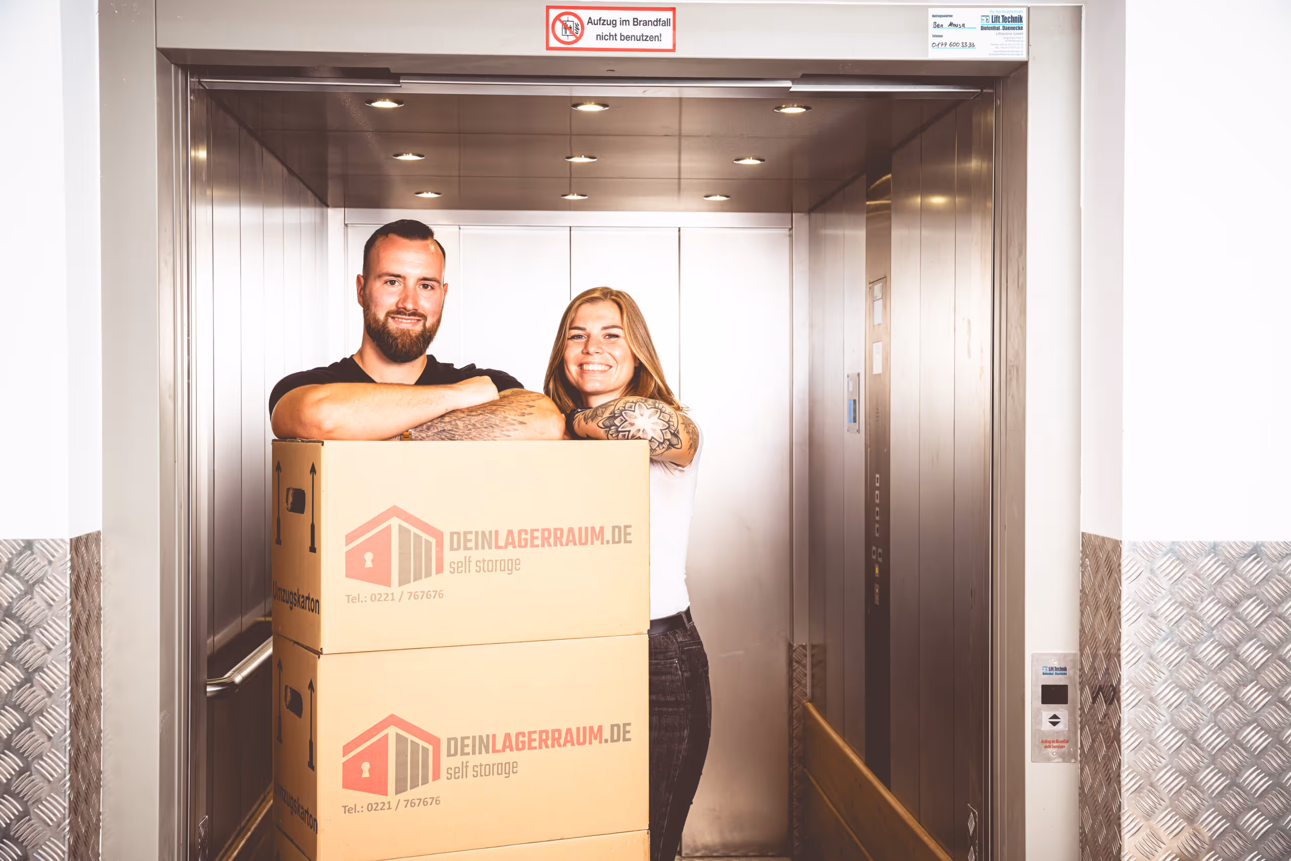 Ein lächelndes Paar steht in einem Aufzug hinter zwei großen Umzugskartons mit der Aufschrift 'DEINLAGERRAUM.DE self storage'.