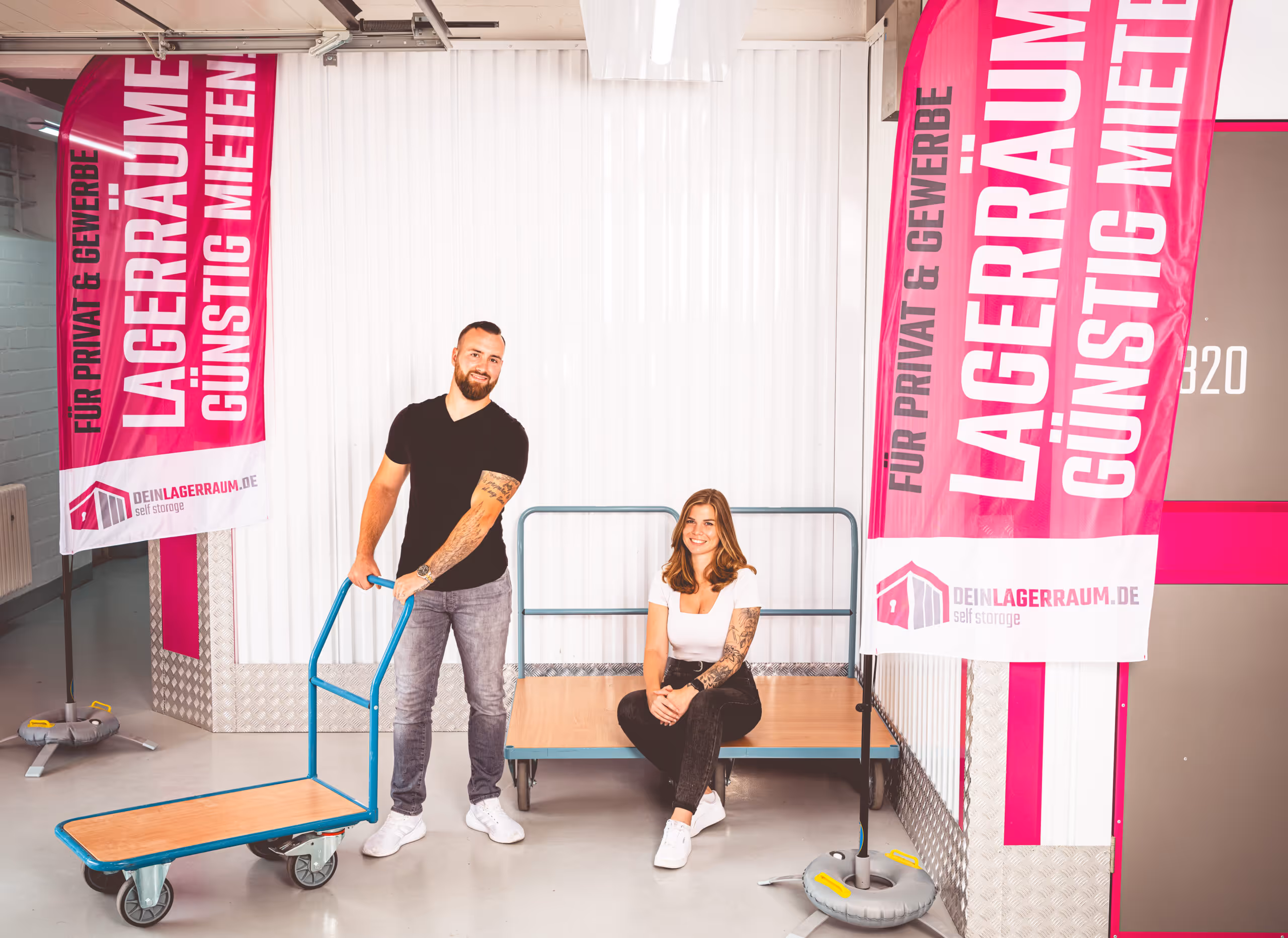 Ein Mann mit schwarzem T-Shirt zieht eine blaue Sackkarre, eine Frau in weißem Oberteil sitzt darauf in einem Lagerraum mit pinkfarbenen Werbefahnen.