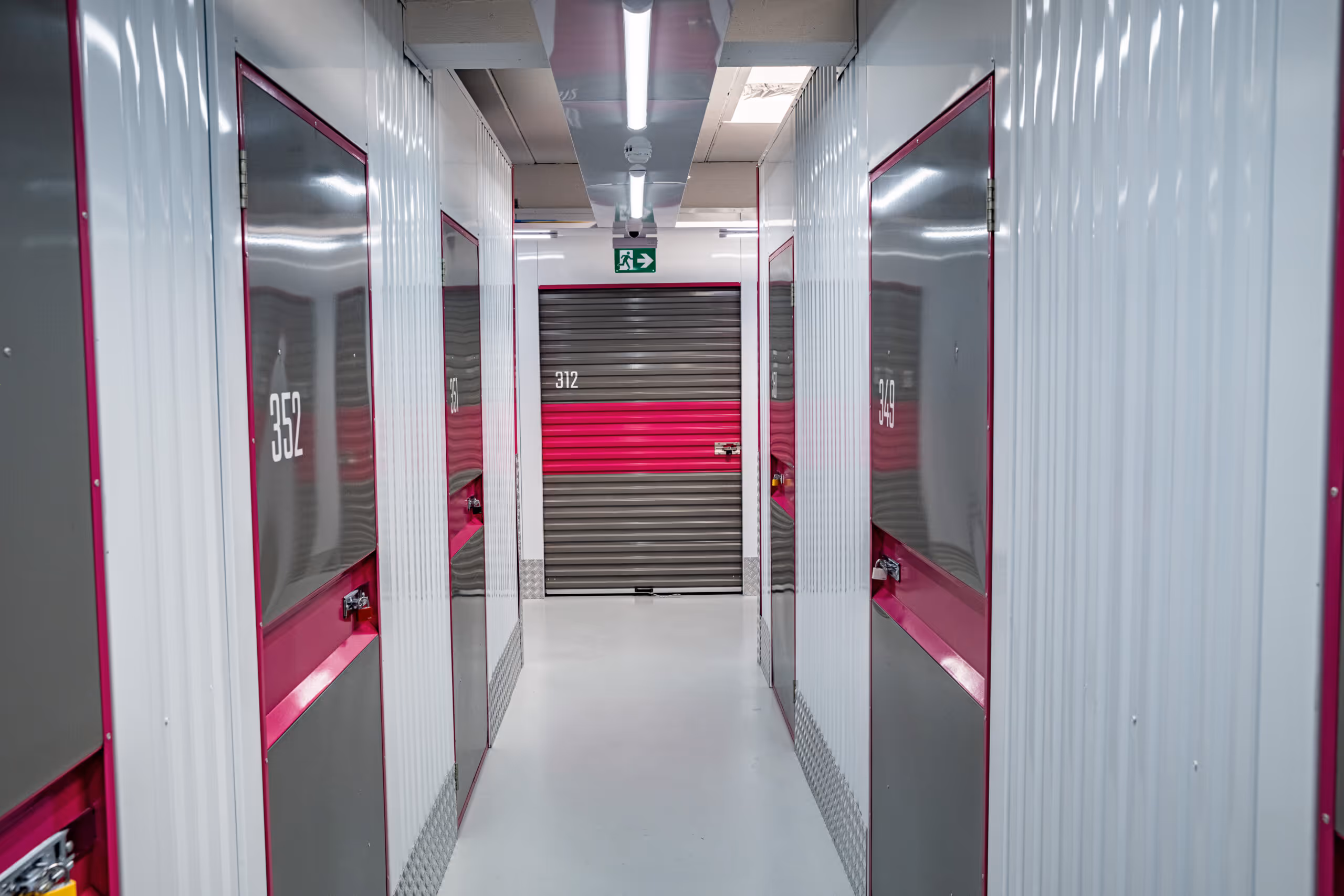 Gang mit verschlossenen Metalllagerboxen in Weiß und Pink, durchgehend beleuchtet mit Fluchtweg-Schild.