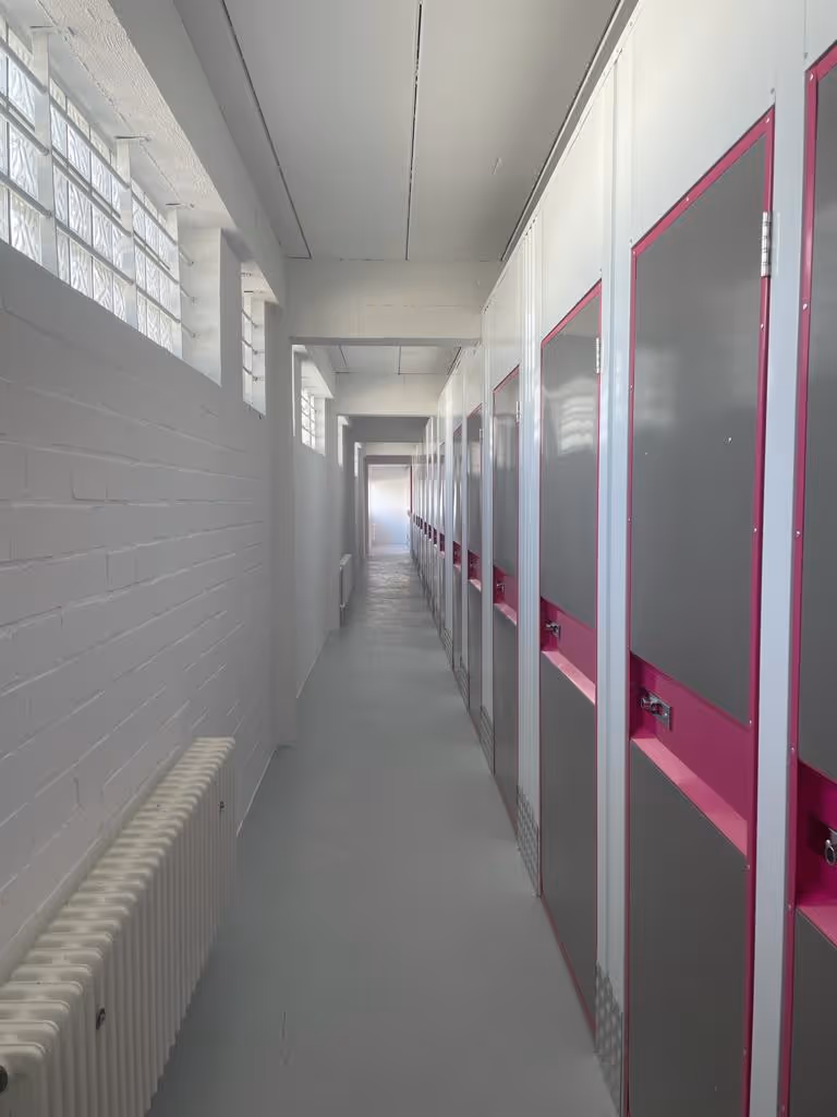 Langer weißer Flur mit grauen und pinkfarbenen Umkleidespinden auf der rechten Seite und Fenstern mit Glasbausteinen links.