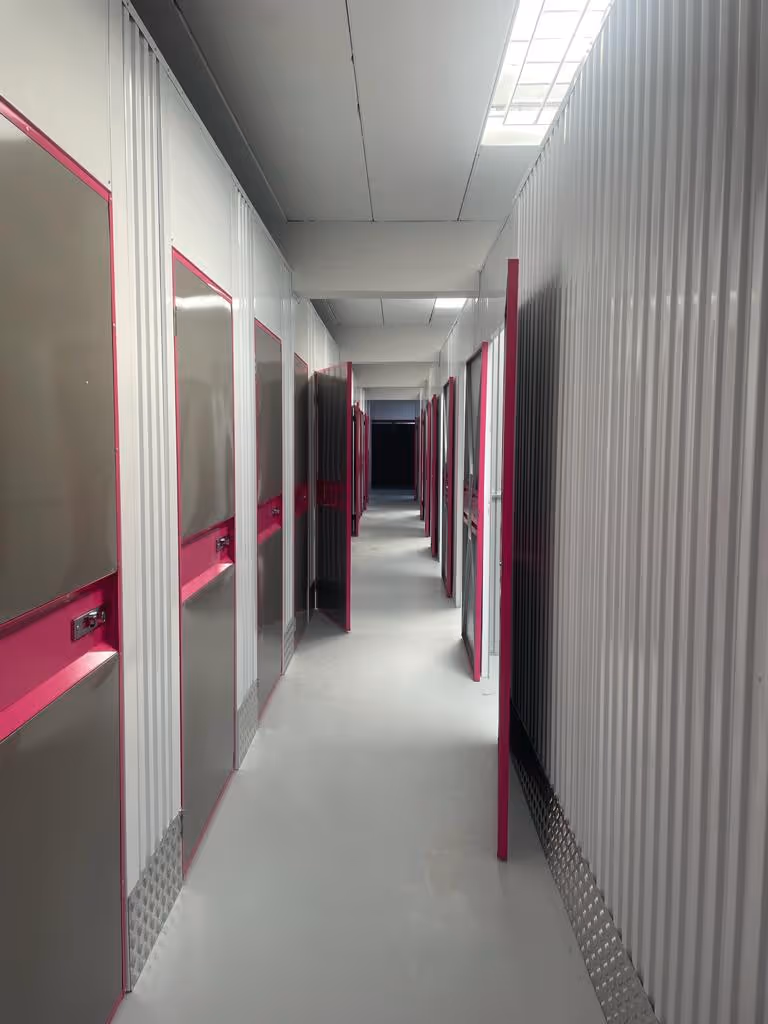 Heller Flur mit mehreren weißen und pink umrandeten Metalltüren, einige davon sind geöffnet.