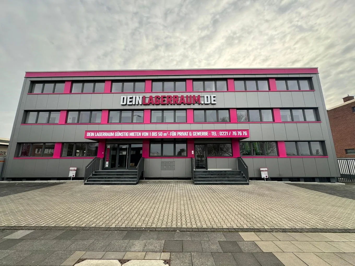Modernes Geschäftsgebäude mit grauer Fassade und pinkfarbenen Akzenten, Schriftzug 'DEIN LAGERRAUM.DE' und Werbebanner für Lagerraumvermietung.
