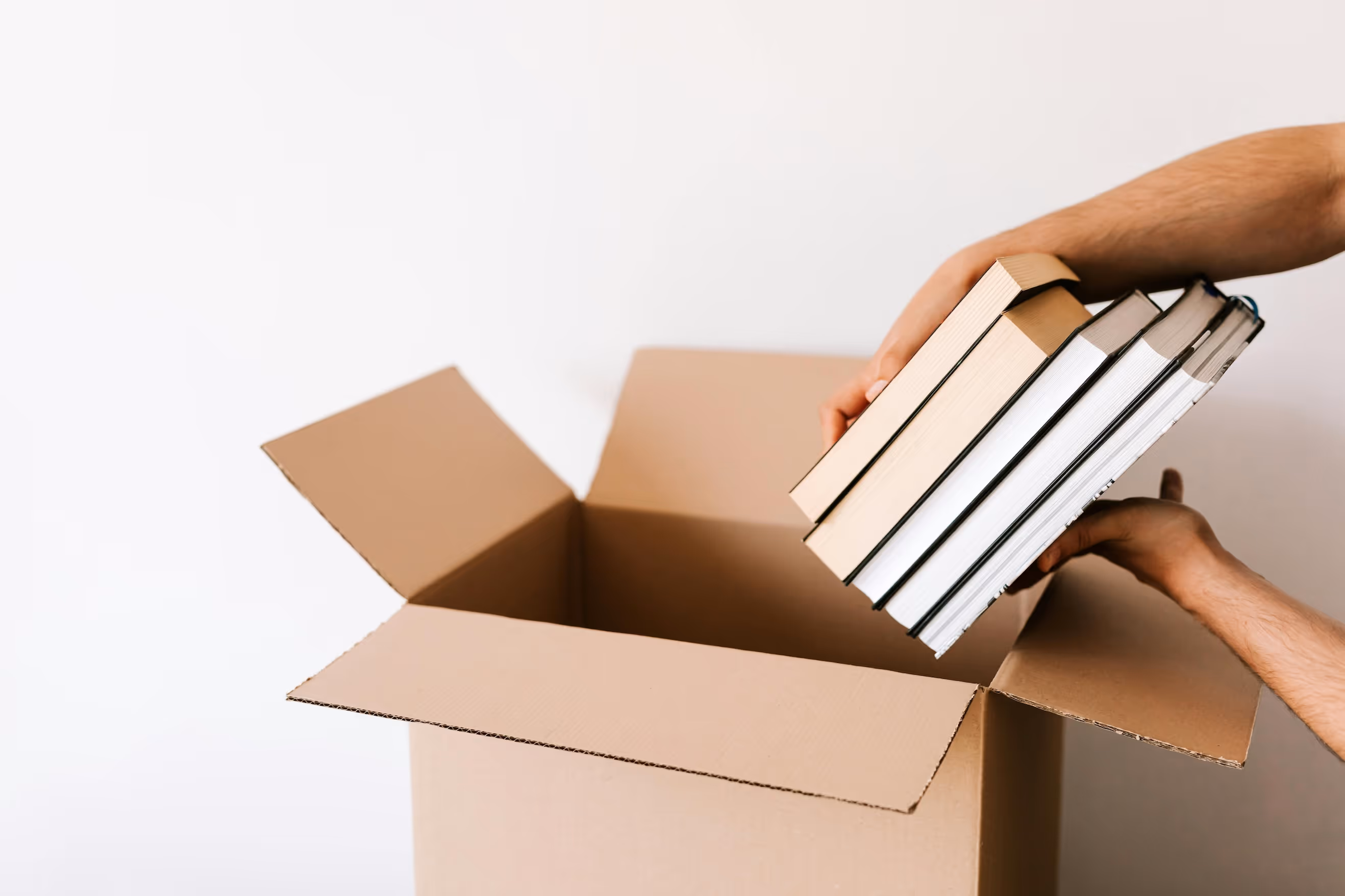 Hände packen fünf Bücher in einen offenen braunen Karton vor weißem Hintergrund.