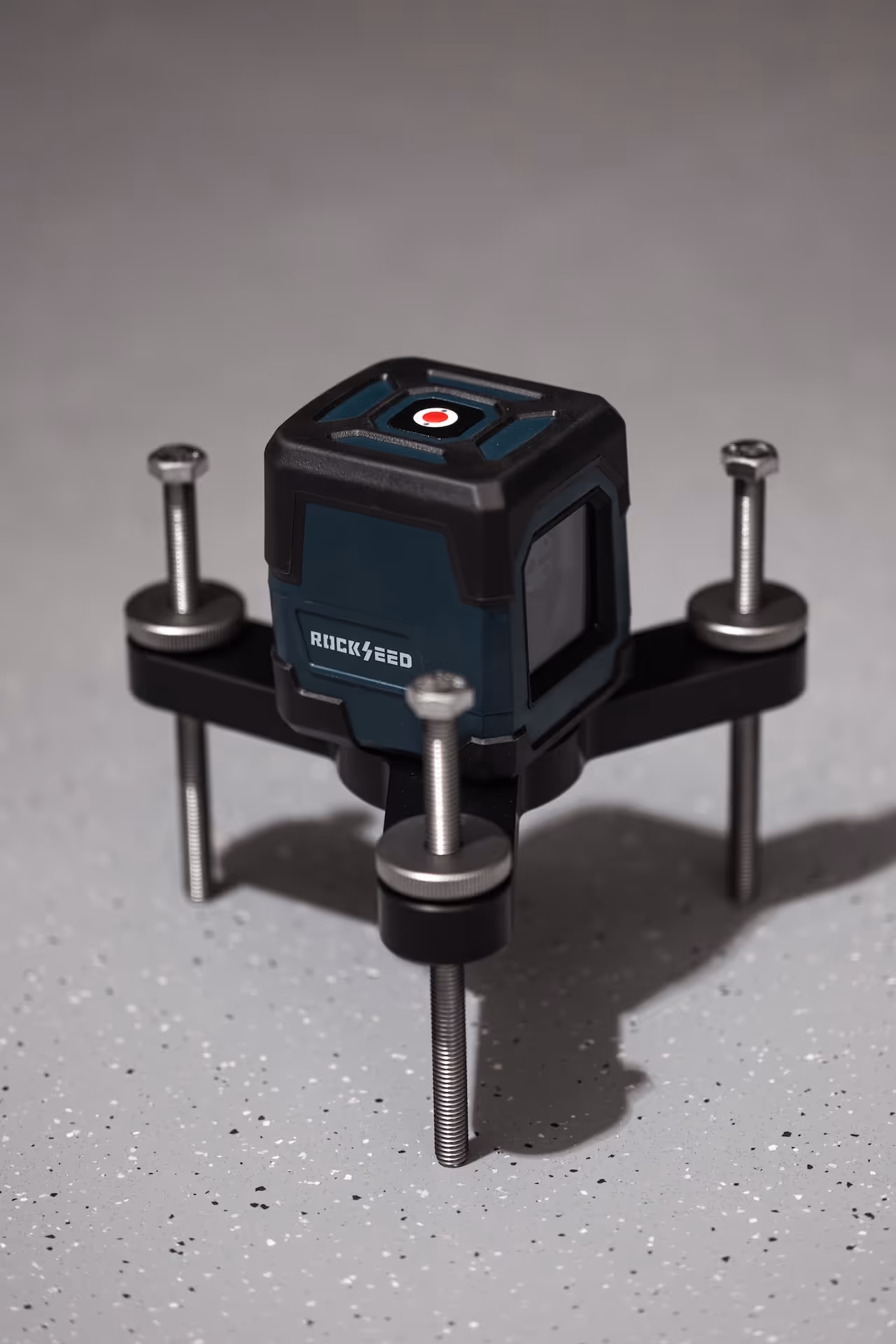 Ricksfeed branded tabletop laser level device with adjustable metal legs on a speckled gray surface.