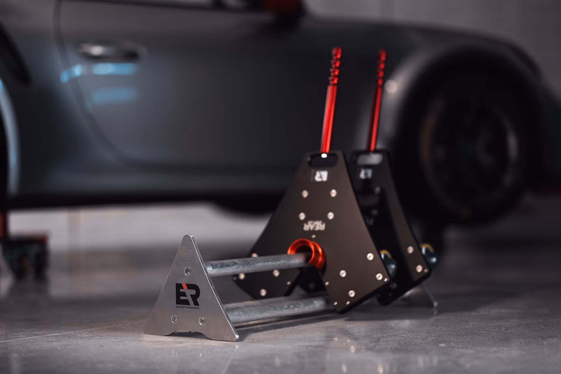 Close-up of a metal car wheel alignment tool with red adjustment handles on a polished floor, with a blurred car in the background.