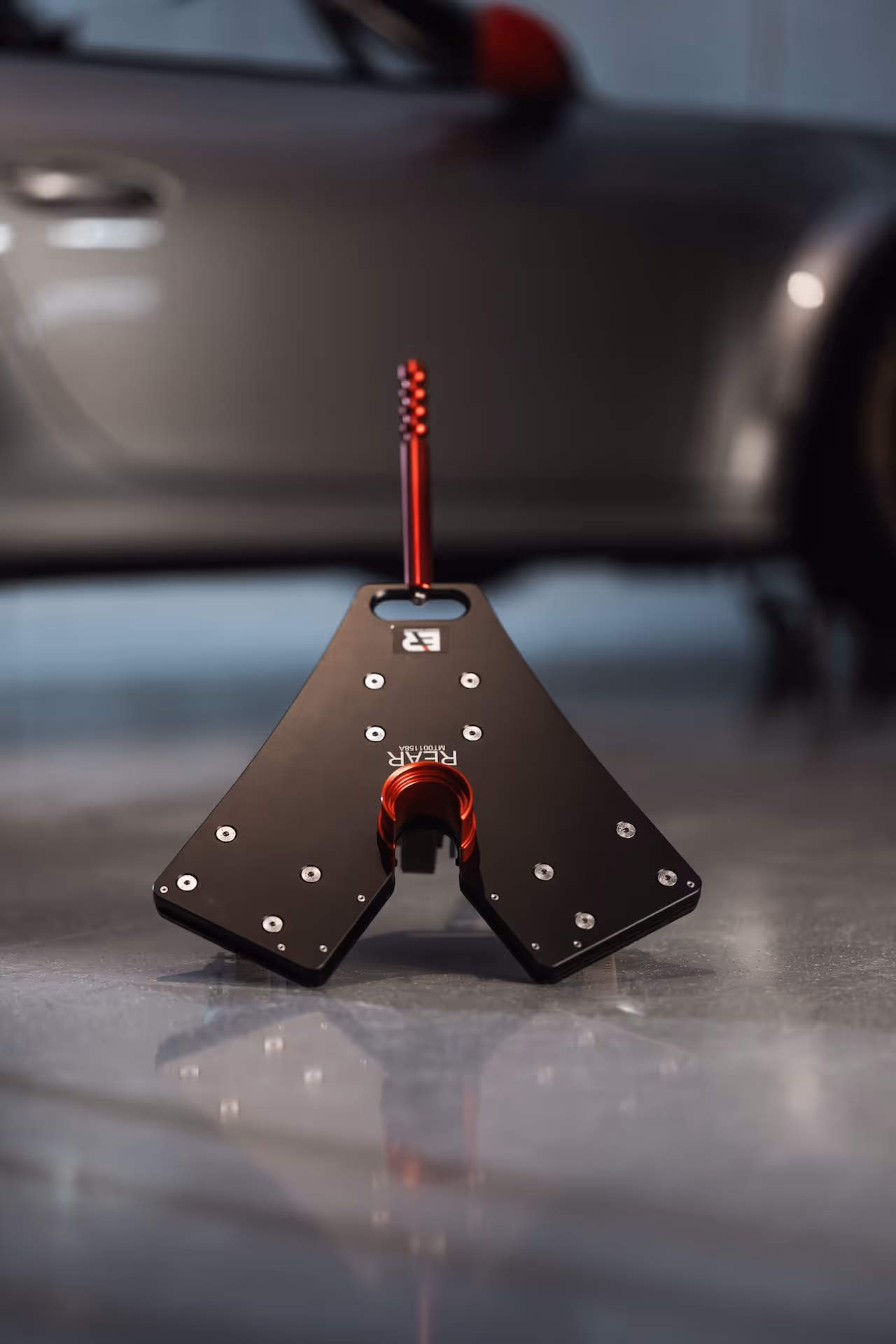 Black metal automotive rear alignment tool with a red handle on a garage floor with a blurred car in the background.
