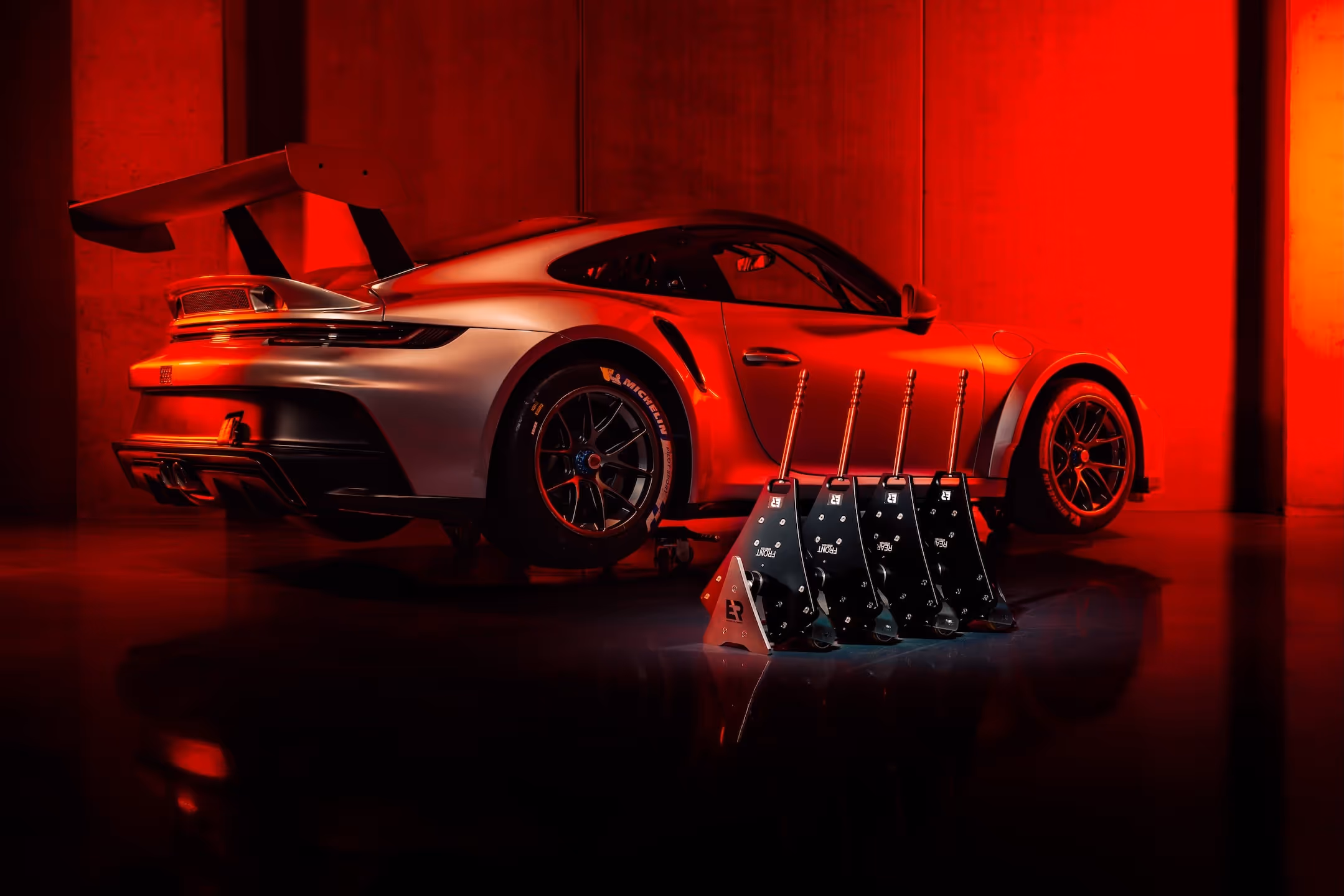Porsche GT3 racecar with a large rear spoiler illuminated by red light, next to a row of five black racing pedal units.