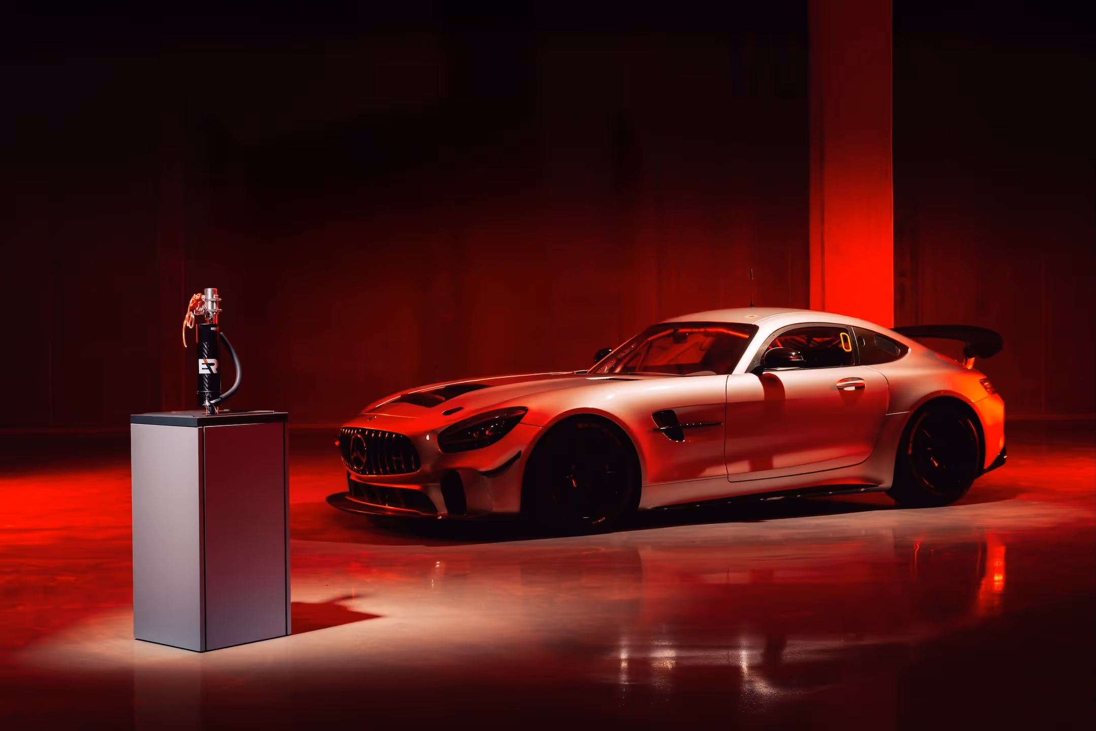 Mercedes AMG GT3 racecar with black accents displayed in a dark room illuminated by red lighting, next to a tall pedestal displaying the lightweight drinking system