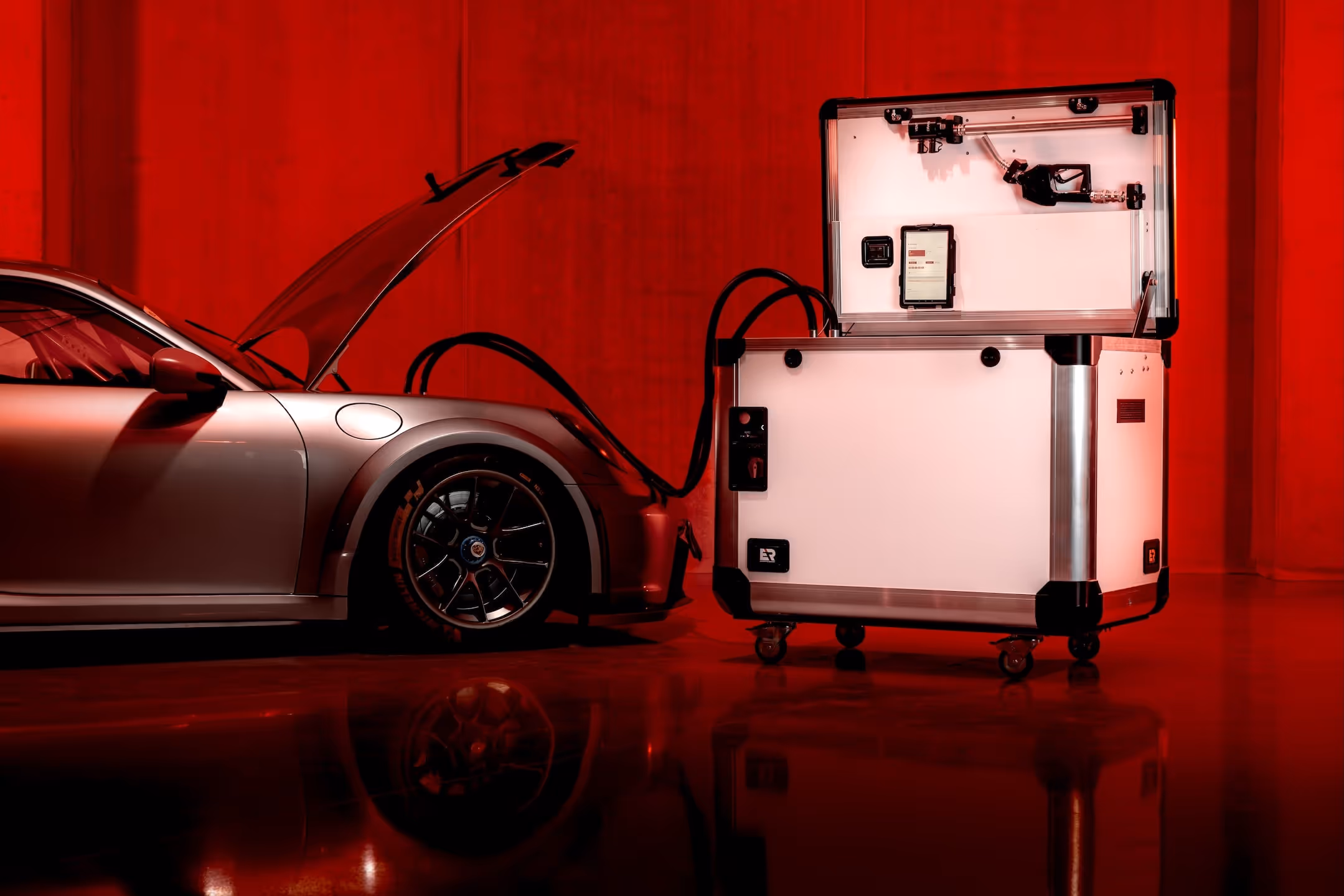 Silver Porsche GT3 racecar with open hood connected via hoses to a large white fuel system in a red-lit room.