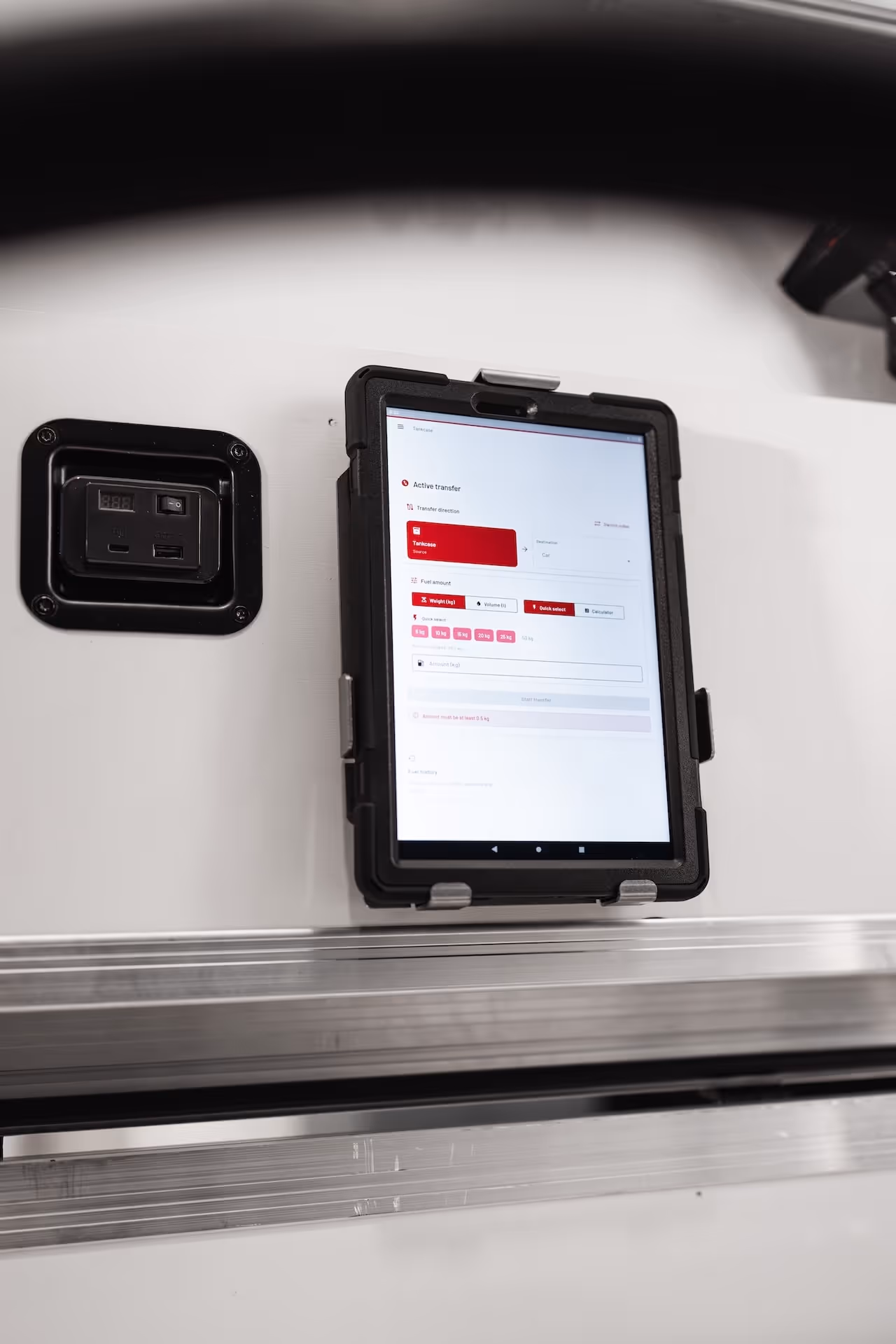 Tablet in rugged case mounted on a white wall displaying an active transfer interface with fuel amount options.